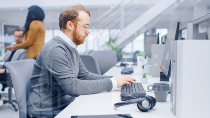 Aufkleber - Young Happy Business Salesman Specialist Works with His Desktop Computer in Bright Modern Office. In the Background Diverse Team of Professional Businesspeople Working