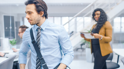 Poster - Young Happy Businessman Walks Into Bright Modern Office, Takes Place at His Desktop Computer. In the Background Diverse Team of Professional Businesspeople Working