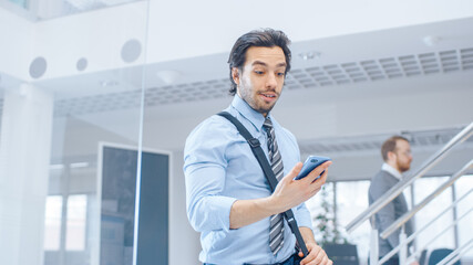 Wall Mural - Young Happy Businessman Walks Into Bright Modern Office, Holds Smartphone and Checks Social Media. In the Background Diverse Team of Professional Businesspeople Working