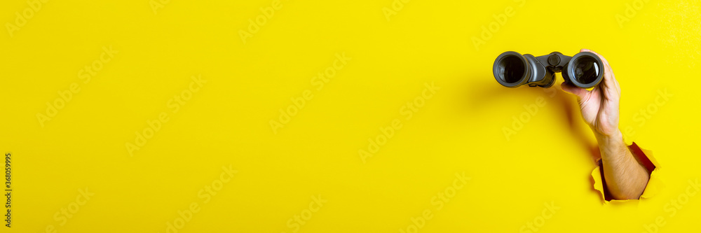 Female hand holds black binoculars on yellow background, travel, find and search concept. Banner - obrazy, fototapety, plakaty 