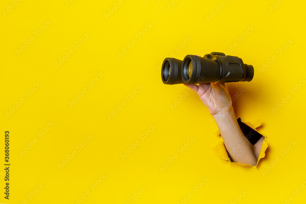 Female hand holds black binoculars on a yellow background. Looking through binoculars, journey, find and search concept - obrazy, fototapety, plakaty 