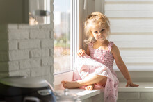 Beautiful Little Girl 4 Years Old In A Pink Dress Sits At The Window On A Sunny Day And Smiles. Sun Glare