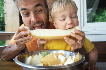 Family eating fresh sweet melon on terrace. Father and his toddler son biting together slice of honeymelon fruit. Summer food.