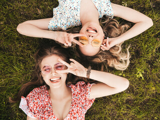 Two young beautiful smiling hipster girls in trendy summer sundress.Sexy carefree women lying on the green grass in sunglasses.Positive models having fun.Top view.They show peace sign