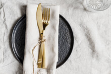Golden Cutlery On A Linen Napkin With Black Plate. Decorated Table Setting For Dinner.