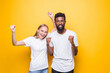 © dianagrytsku - We are the champions. Joyful interracial couple rejoicing success, raising fists, celebrating victory, posing together over yellow background