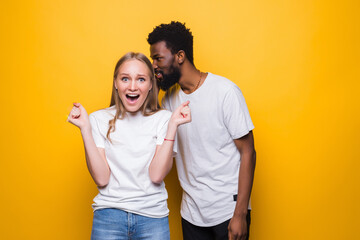 Cheerful young mixed couple whispering secret behind her hand sharing news posing isolated on yellow background. People lifestyle concept.