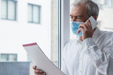 Wall Mural - mature man with medical mask using mobile phone and flipping through documents in office, new normal concept