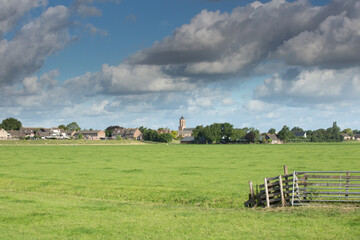 Summer pasture landscape with fresh green grass and checkered fence with village view in the background with Kerk de Woudse Dom in Rijnzaterwoude against sky with blue spots and dark gray clouds