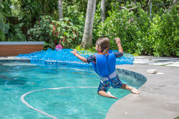 Child boy jumping in the air stop action into a pool at home backyard on a sunny day of summer in Miami Florida wearing life vest blue net in background