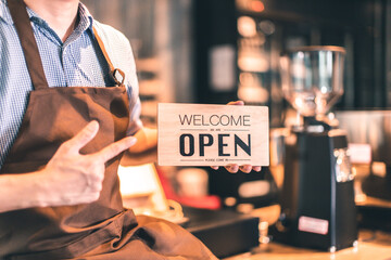 Business owner attractive young Asian man in apron hanging we're open sign on front door  welcoming clients to new cafe. Happy waiter with protective face mask holding open sign while stand at cafe .