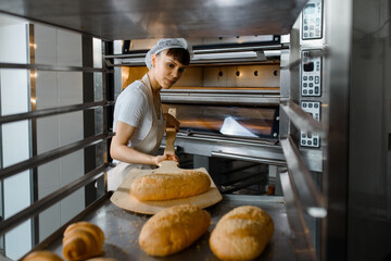 Wall Mural - Close up of young caucasian woman baker putting the fresh bread on the shelves/rack at baking manufacture factory.