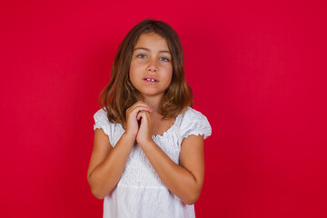 Positive adorable Little caucasian girl with blue eyes wearing white dress standing  smiles happily, glad to receive pleasant news from interlocutor, keeps hands together. People emotions concept.