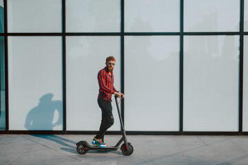 A handsome young bearded businessman rides  e scooter in front of the white wall of the building