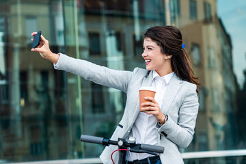 Wall Mural - Businesswoman with electric scooter standing in front of modern business building taking selfie with phone.