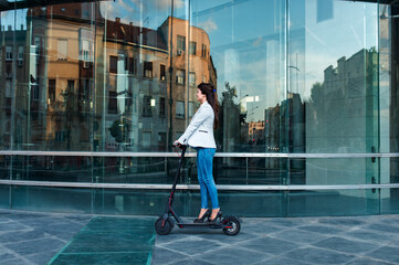 Wall Mural - Young businesswoman driving electric scooter in front of modern business building.