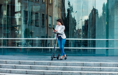 Wall Mural - Businesswoman with electric scooter standing in front of modern business building looking at phone.	