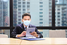 Asian Male Employee In Formal Suit Wearing Protective Facial Mask Working Online With Tablet In Business Office During Coronavirus Or Covid-19 Pandemic As New Normal And Social Distancing Policy