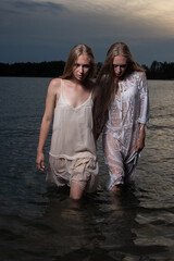 two attractive young twin sisters with long blond hair posing in light dresses in water of lake at summer night. stylish fashion photoshoot with flashlight. pretty models outdoors evening photosession