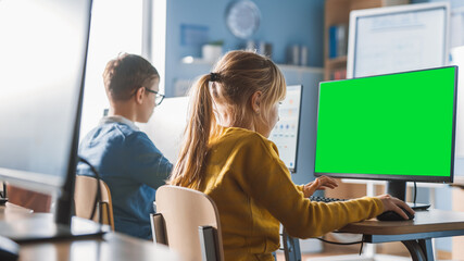 Elementary School Computer Science Classroom: Cute Little Girl Uses Green Mock-up Screen Computer while Learning Coding and Programming. Schoolchildren Getting Modern Education. Over the Shoulder