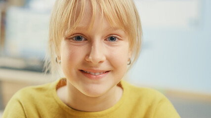 Portrait of a Cute Little Girl with Blond Hair Sitting at her School Desk, Smiles Happily. Smart Little Girl with Charming Smile Sitting in the Classroom. Close-up Shot