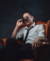 Young confident man in sunglasses wearing white shirt and black tie sitting on a luxury chair and looking on the camera against black background