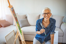 Portrait Of Attractive Middle Aged Professional Female Painter With Gray Hair Working At Home Standing In Front Of Easel. Happy Mature Woman Painting And Watching At Camera.