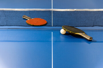 Black and red table tennis racket and a white ball on the blue ping pong table with a net, Two table tennis paddle is a sports competition equipment indoor activity and exercise for background concept