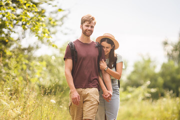 Caucasian hipster couple walking along path in countryside. man with beard holds the hand of his beloved girl while hiking in natural park with backpack. Travel, hiking, tourism and people concept