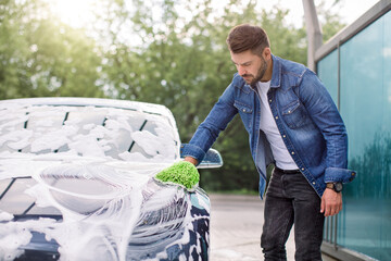 Wall Mural - Outdoor car wash concept. Portrait of young man in casual wear, washing his luxury modern car in a self-service car wash station with sponge and foam.