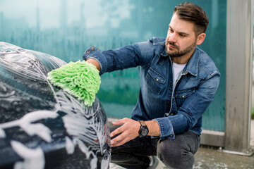 Wall Mural - Young handsome Caucasian man cleaning his luxury electric car headlights outdoors at self car wash station, using green rag mitten and soap foam.