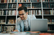 © Dusko - Young nice looking student or writer sitting in college library next to the window and working on his laptop. Natural light with strong and deep shadows. Wall with books in the background.