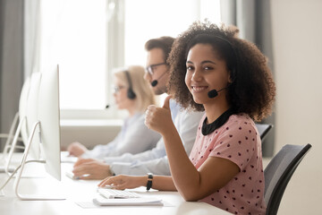 Wall Mural - smiling mixed race female call center operator, agent in wireless headset showing thumbs up gesture,