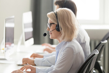 Wall Mural - mature female call center operator in wireless headset consulting client online, working on computer