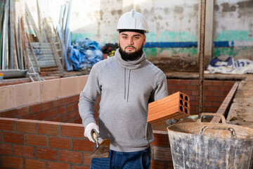 Wall Mural - Young bearded bricklayer installing brick wall in building under construction..