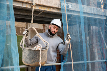 Wall Mural - Confident bearded guy in construction helmet working on his house renovation, lowering down bucket with tools on rope..