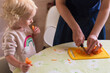 © karinabost - Small girl with blond curly hair supporting her mother cooking in the kitchen. Mother is cutting carrot and the girl is looking and eating slices. Healthy eating and family time concept.