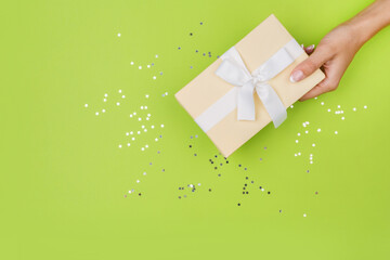 Women hands holding a gift or gift box decorated with confetti on a green table top view. Flat composition for birthday or wedding.