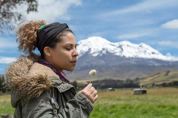 Portrait of a beautiful latina girl look and blow a dandelion flower she wears a heavy coat and in the background the highest mountain in Ecuador the Chimborazo volcano