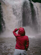 A woman wearing a red hood, standing with her back turned. Blur backdrop is wonderful waterfall in the rainy season, Thailand. Feeling fresh, relaxed, and freedom. Ideas for adventure nature tourism.