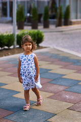 A little girl in a dress stands on the playground and the wind develops her hair. The child is very serious