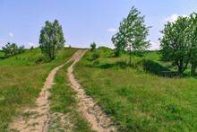 Two-lane Dirt Road Up A Hill In The Middle Of Meadows And Lush Green Grass On A Sunny Spring-summer Day. Natural Landscape With A Path Over The Horizon And A Wooden Table/bench In The Shadow Of A Tree