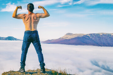 young man of athletic build stands on top of a mountain with clouds demonstrating the strength and power of a man