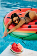 © shevtsovy - sexy woman relax in swimming pool on inflatable pool toy watermelon outdoors at hot summer day. Sexy female model in swimsuit lay on inflatable mattress watermelon in swimming pool outdoors