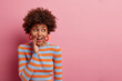 Surprised curious curly woman keeps hand near mouth and whispers secret, spreads rumors, looks with wondered expression aside, dressed casually, isolated on pink background, blank empty space