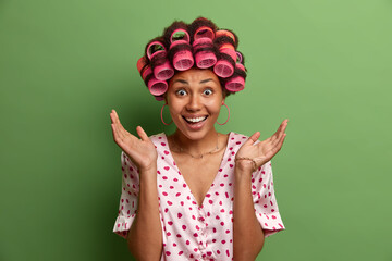 Joyful dark skinned woman spreads palms, has happy reaction on hearing awesome news, wears hair rollers, casual silk dressing gown, poses against green background, prepares for special meeting