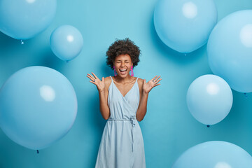 Wall Mural - Shot of upbeat cheerful dark skinned woman feels very happy and excited, raises palms and laughs, spends free time on party, wears nice blue summer dress with earrings and rings, poses near balloons