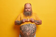 © wayhome.studio  - Serious bearded man with big tummy, tattooed arms and belly, holds very small carton cup of coffee containing much sugar, enjoys caffeine aromatic beverage, wears yellow t shirt, poses indoor alone