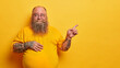 © wayhome.studio  - Smiling fat bearded man in eyewear with bald patch and tattooed arms, points on empty space aside, has big stomach after drinking beer, promots something on yellow background, recommends product
