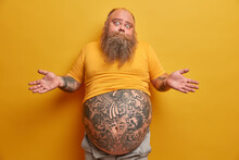 Hesitant Thick Man With Big Tattooed Belly, Shrugs Shoulders And Looks Confused, Faces Dilemma, Makes Serious Decision, Wears Undersized Yellow T Shirt, Poses Indoor. People And Doubt Concept